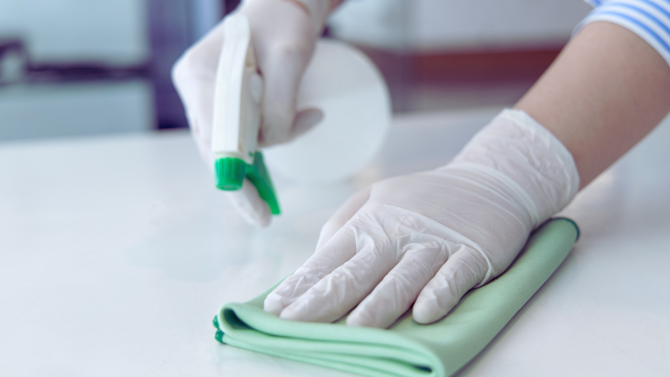 What You Need to Know About Surface Sanitization for a Safer Living Space Person wearing gloves using a spray bottle and cloth for surface sanitization, emphasizing safer living space through proper cleaning practices.